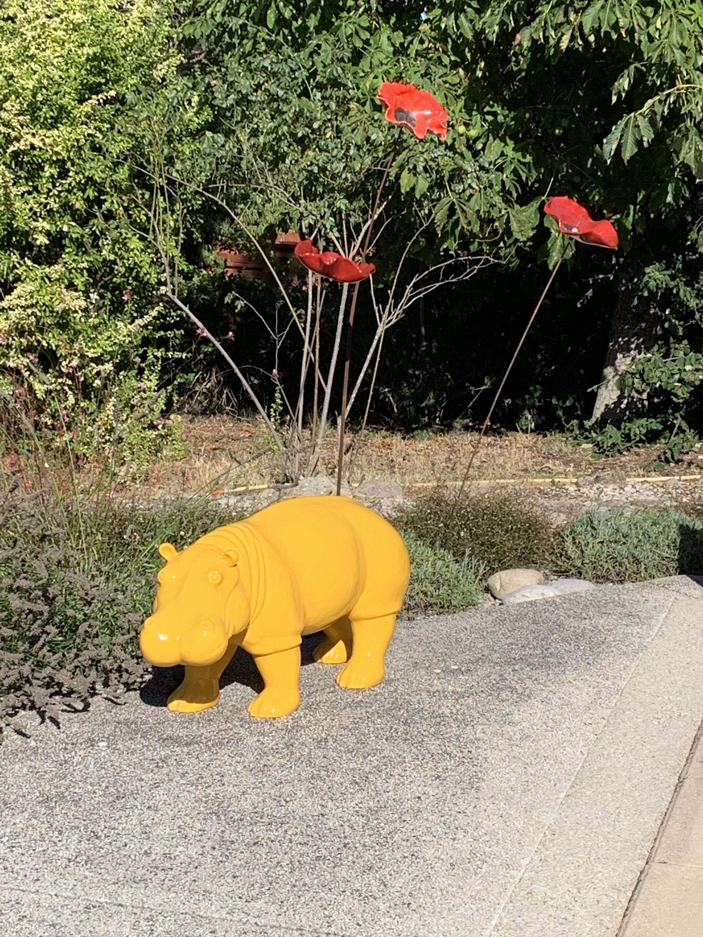 A yellow plastic hippopotamus sculpture on a paved surface with red flowers on a nearby plant.