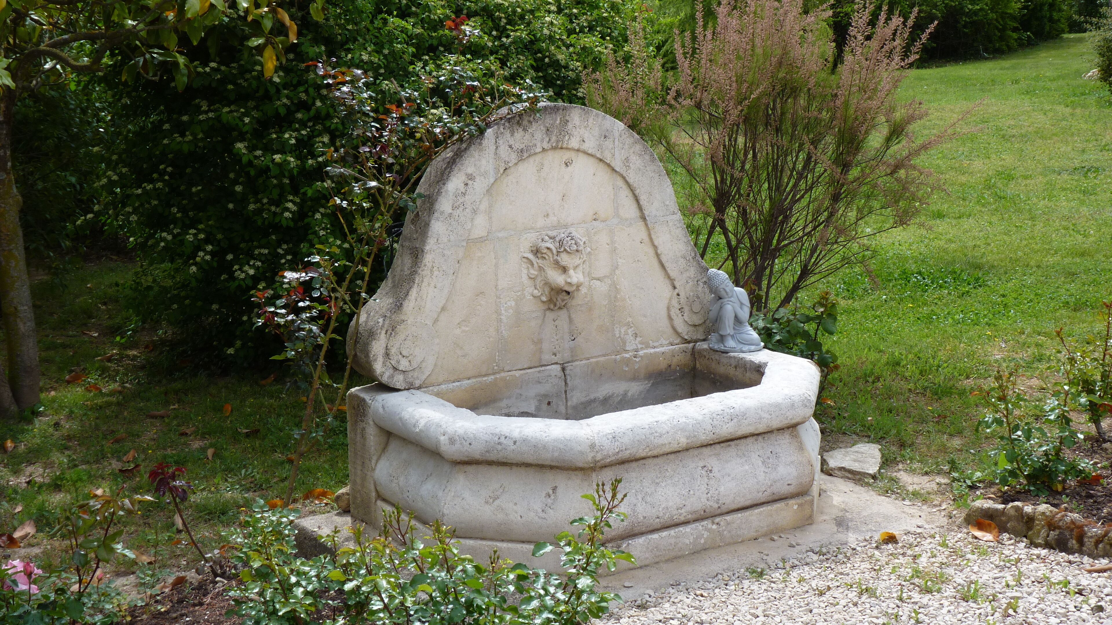 A stone fountain with a sculpted figure, surrounded by greenery and a grassy area.