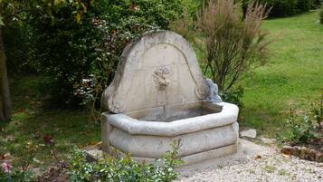 A stone fountain with a sculpted figure, surrounded by greenery and a grassy area.