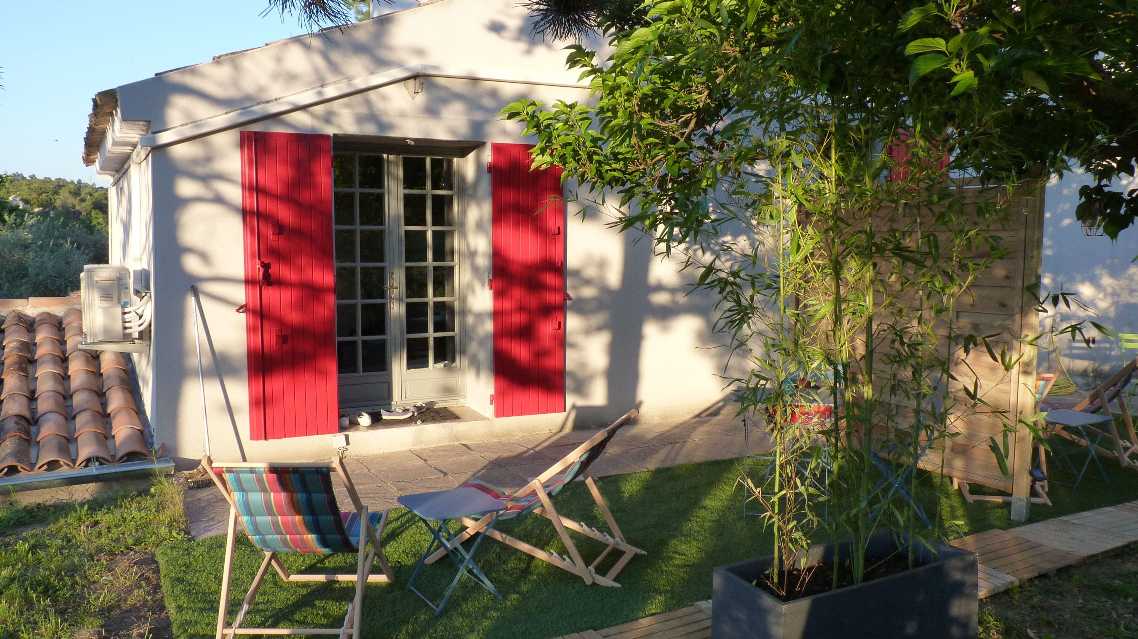 Een huis met rode luiken, een klein voortuintje en een tuin met een boom en twee stoelen.