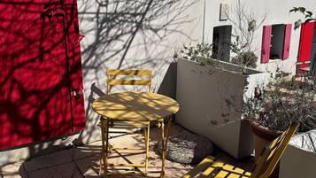 Un patio con una silla amarilla y una mesa, un portón rojo y un edificio con contraventanas rojas al fondo.