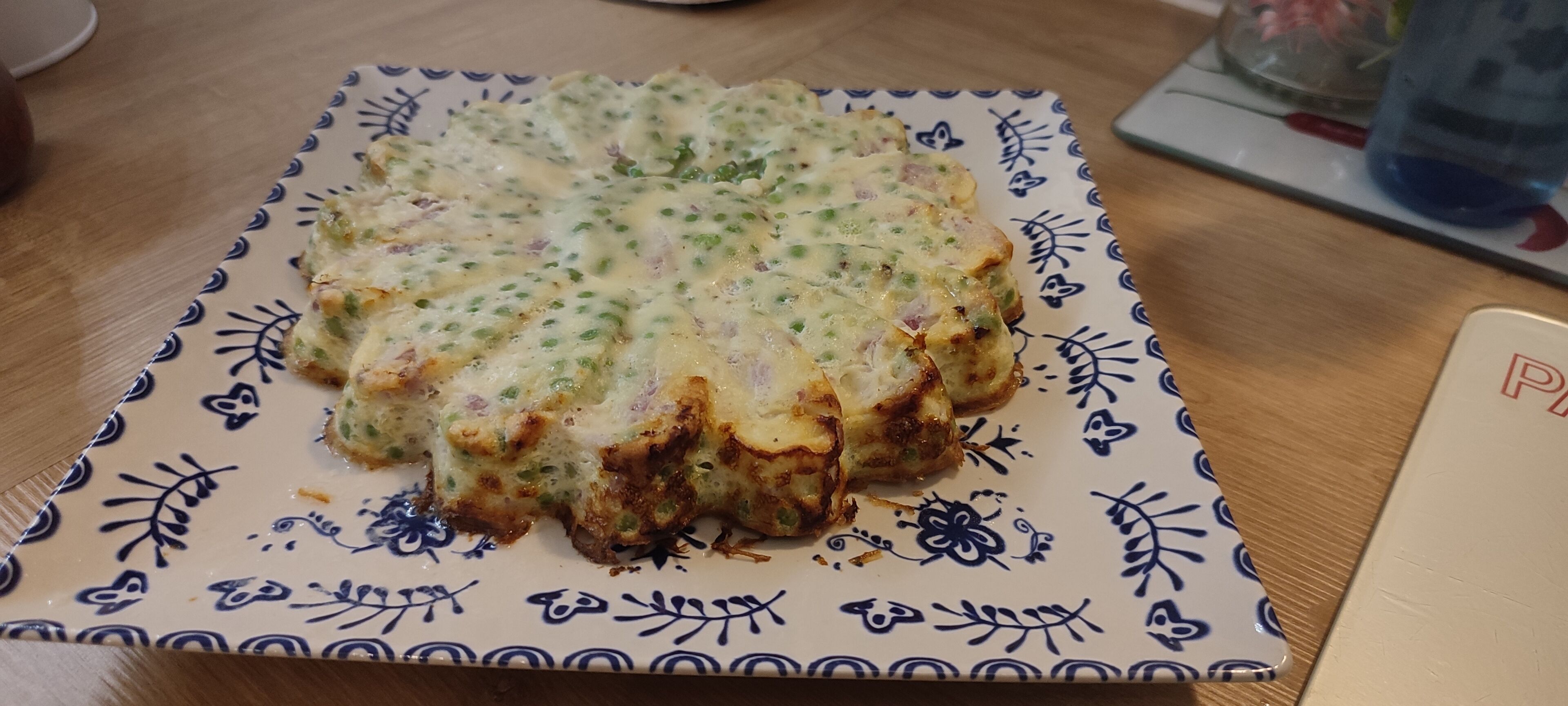 A square plate with a floral pattern, containing a food item with green specks, possibly a vegetable or herb, on a wooden table.