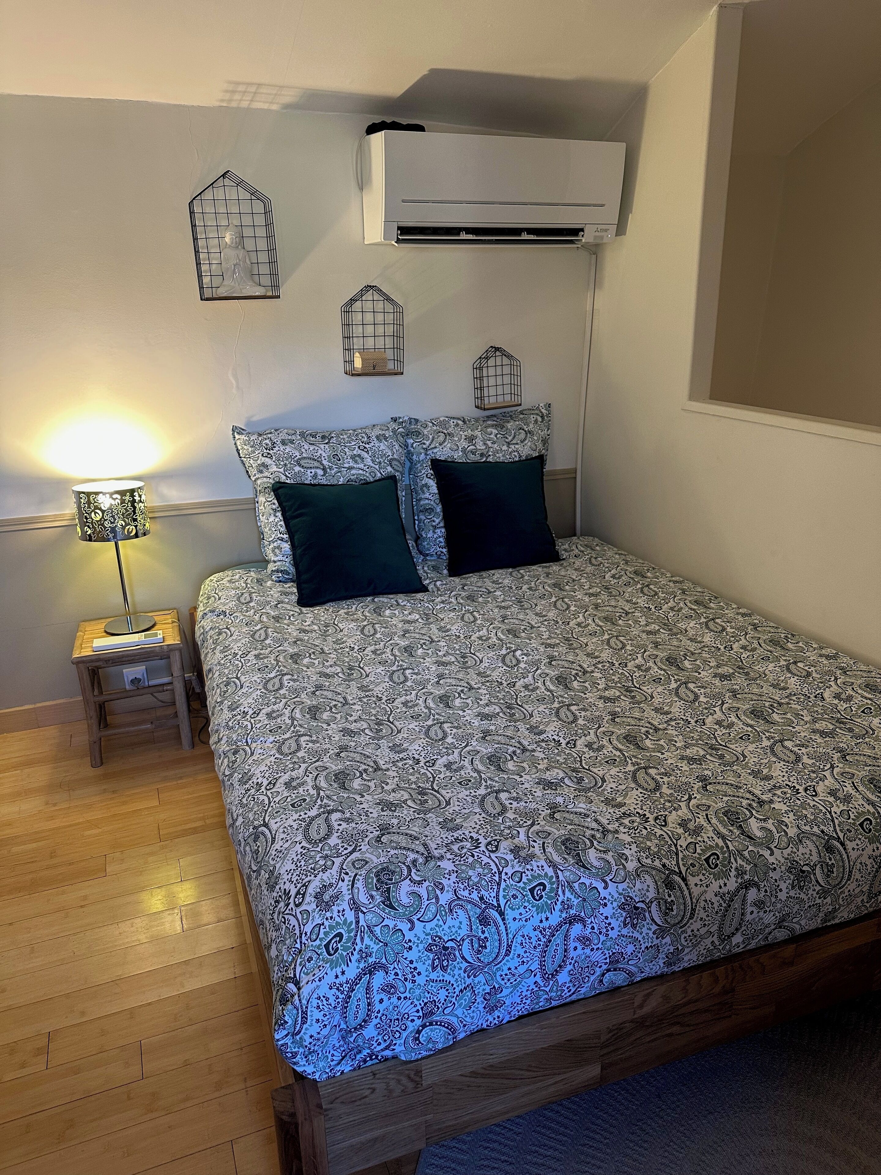 A bedroom with a patterned bedspread, two pillows, a bedside lamp, a small wooden nightstand, and an air conditioning unit mounted on the wall.