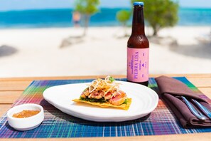 Plato con comida, una botella de cerveza Piel de Perla y una servilleta sobre una mesa con un mantel individual colorido.