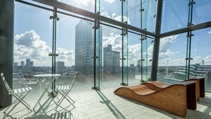 A modern interior with a panoramic city view, a glass partition, and a wooden bench. - City Suite - Rooftop Spa - Balcony - Skyline View (Manchester)
