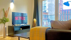 A modern living room with a flat-screen TV displaying 'NETFLIX', a sofa, a coffee table, a potted plant, and a large window with a view of buildings. - City Suite - Rooftop Spa - Balcony - Skyline View (Manchester)