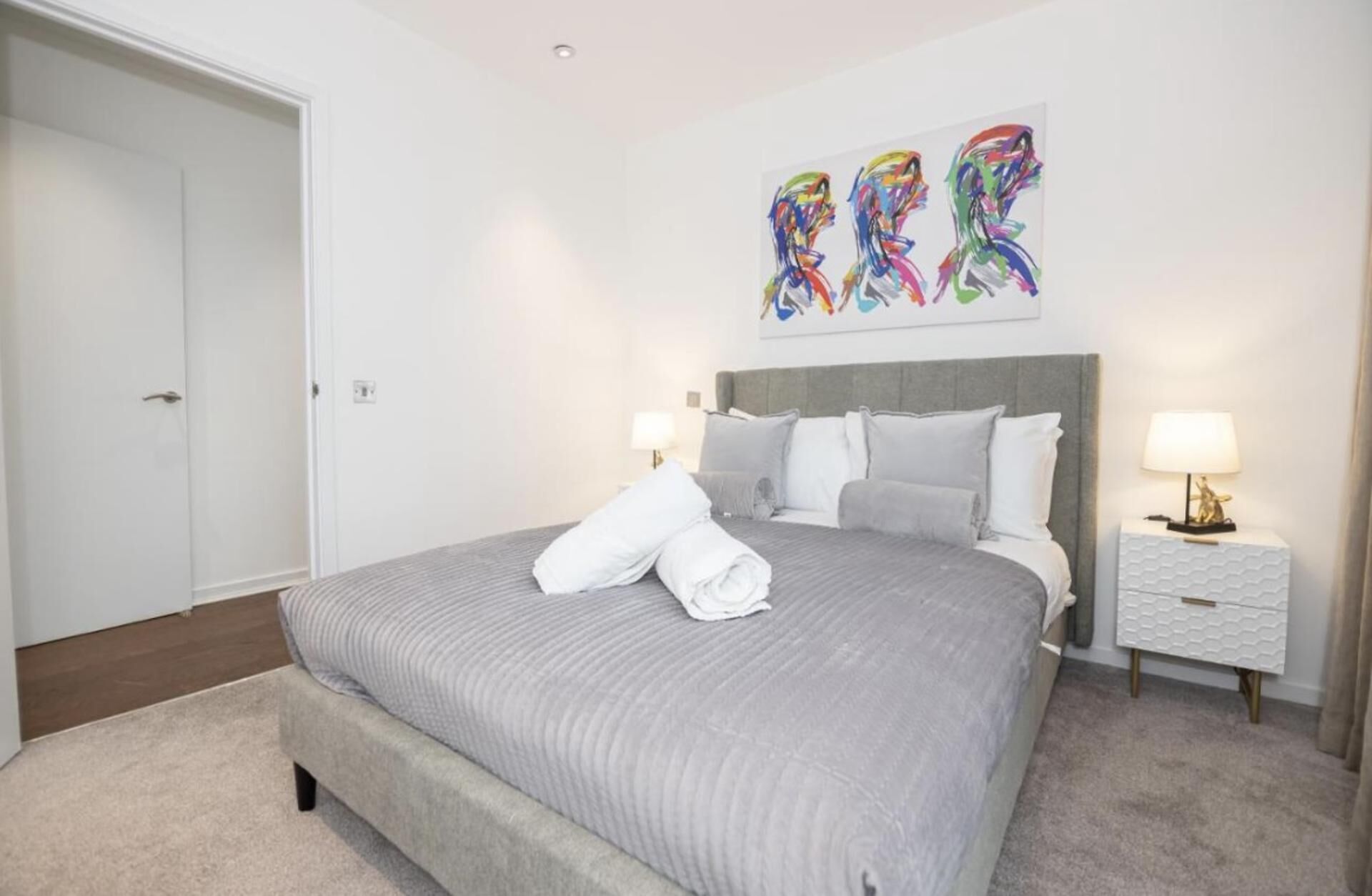 Uma cama bem arrumada com manta e almofadas cinzas, cabeceira cinza, mesa de cabeceira branca com uma luminária, e uma pintura abstrata colorida acima da cama.