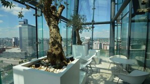 A modern interior with a large tree planter, seating area, and panoramic city views through large windows. - City Suite - Rooftop Spa - Balcony - Skyline View (Manchester)