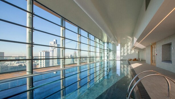 A modern indoor swimming pool with large glass windows, a clear view of the cityscape, and a minimalist design. - City Suite - Rooftop Spa - Balcony - Skyline View (Manchester)