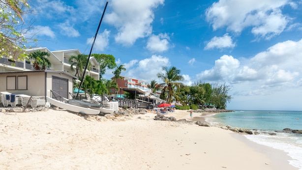 A beach with a building, palm trees, and a clear sky. -  A charming blend of comfort and sophistication in this near-the-beach townhouse (Holetown)