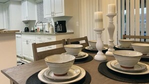 Un comedor con una mesa preparada para cuatro, armarios blancos, una cocina al fondo y una ventana con persianas.