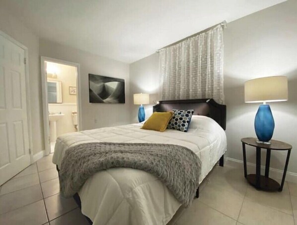 A bedroom with a bed, two bedside lamps, a nightstand, a window with curtains, and a painting on the wall.