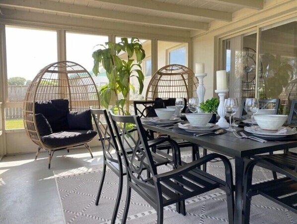 Comedor con una mesa lista para dos personas, rodeada de sillas, una silla colgante y una planta grande en maceta.