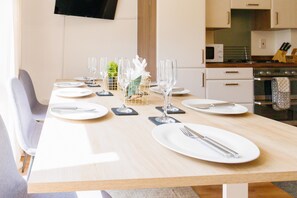 A dining table set for four with white plates, cutlery, and wine glasses, in a modern kitchen with white cabinets and a microwave. - Orchid House in Cambridge (Cambridge)