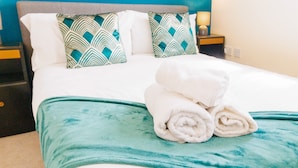 A neatly made bed with white linens and teal pillows, a teal throw blanket, and a stack of white towels on the bed. - Orchid House in Cambridge (Cambridge)