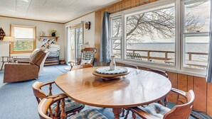 A dining area with a wooden table and chairs, a blue and white patterned rug, and a view of the water through large windows.