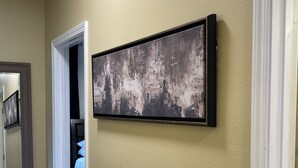Ein Flur mit einem gerahmten abstrakten Gemälde an der Wand, einer zur Schlafzimmers führenden Tür und einem Spiegel, der einen Teil des Flurs reflektiert.
