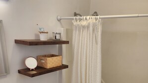 Un baño con inodoro, lavabo y bañera. Hay estantes de madera sobre el inodoro y una cortina de ducha blanca.