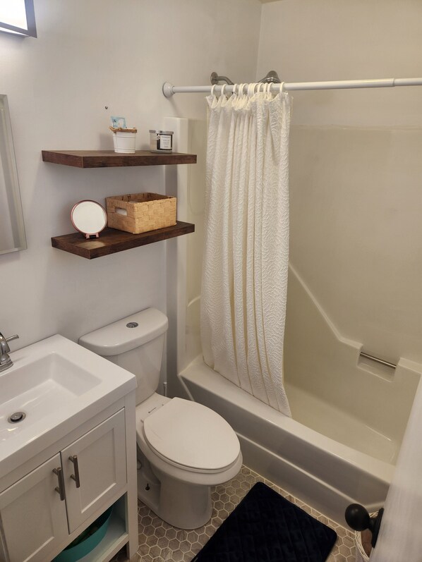 Un baño con inodoro, lavabo y bañera. Hay estantes de madera sobre el inodoro y una cortina de ducha blanca.