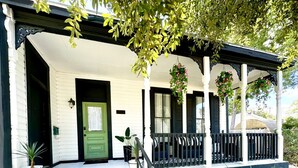Un edificio con puerta verde, paredes blancas y un porche adornado con macetas colgantes de flores.