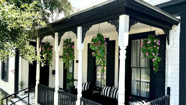 Un porche con columnas blancas, barandillas negras y cestas de flores colgantes.