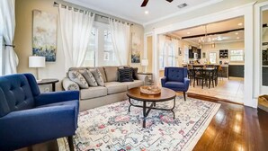 Un salon avec un canapé, deux fauteuils, une table basse et un tapis à motifs. On y trouve également un ventilateur de plafond, un coin repas avec une table et des chaises, et une cuisine à l’arrière-plan.
