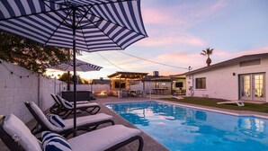 Ein Swimmingpool mit Liegestühlen und Sonnenschirmen, ein Grillplatz sowie ein Haus im Hintergrund.