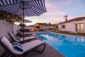 En pool med solstolar och parasoller, ett område med grillplats och ett hus i bakgrunden.