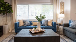 Ein Wohnzimmer mit einem blauen Sofa, einem Couchtisch mit Dekoration, einer Zimmerpflanze und einem Schild mit der Aufschrift 'RELAX'.