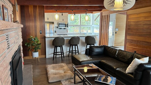 A cozy living area with a leather sofa, a coffee table, and a fireplace. There are three bar stools and a potted plant. The kitchen is visible in the background with white cabinets and a large window.