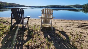 Zwei Adirondack-Stühle, die auf einen ruhigen See mit einem bewaldeten Hügel im Hintergrund blicken.