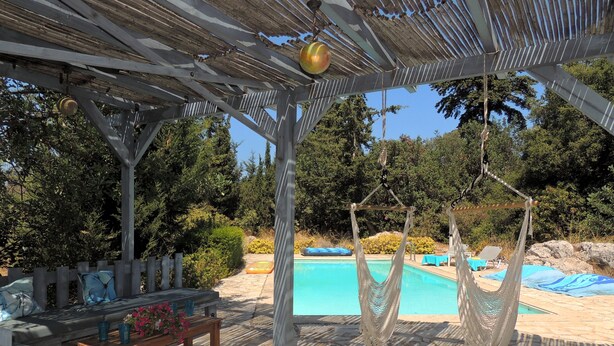Un área junto a la piscina con una pérgola de madera, dos hamacas, un banco con cojines y una mesita con un jarrón de flores.