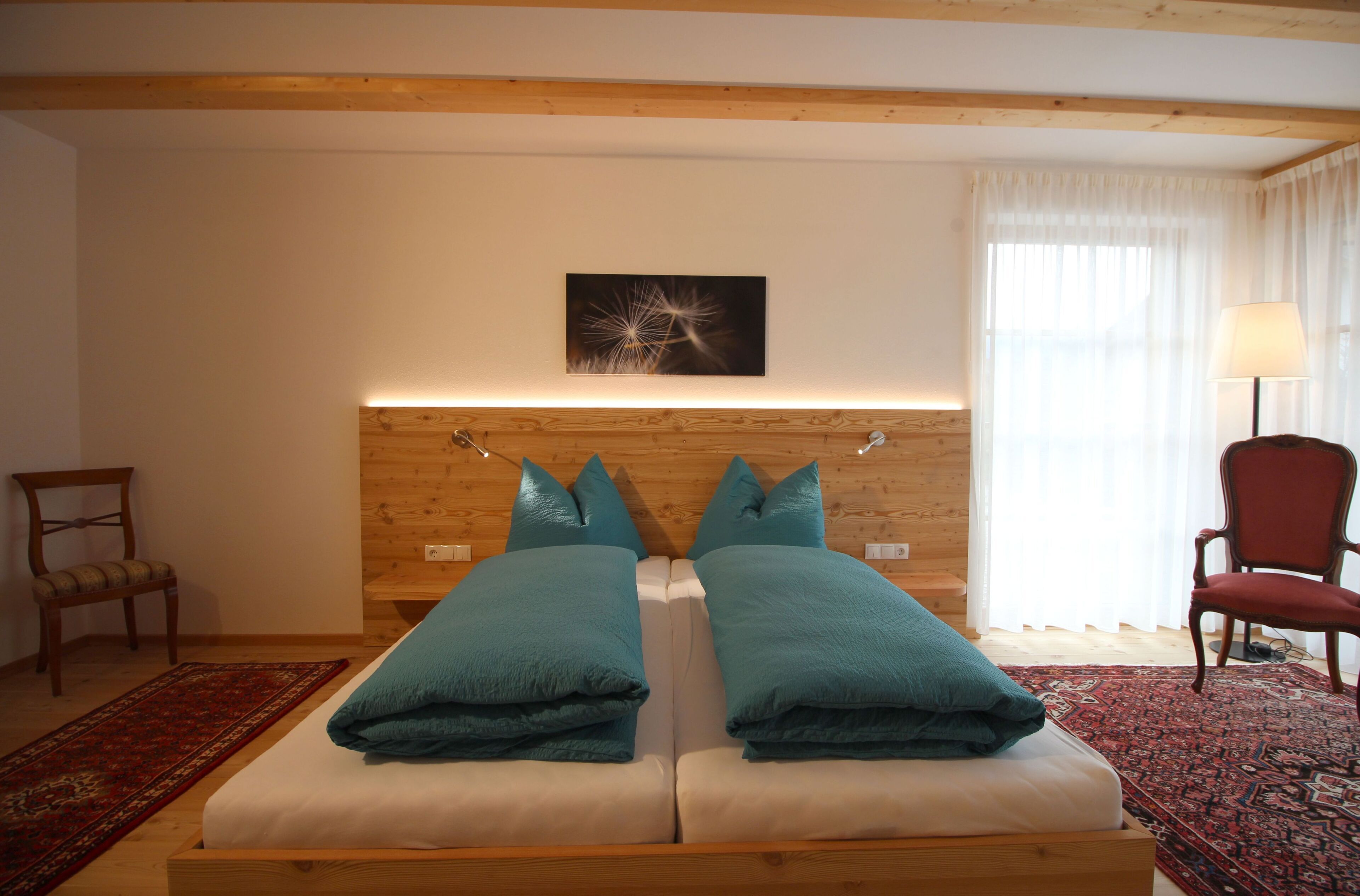 A double bed with teal pillows, a wooden headboard, a red chair, and a red and black patterned rug.