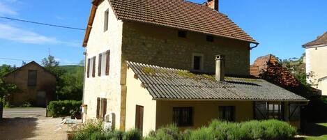 A two-story stone house with a tiled roof, a gravel driveway, and a small garden area.