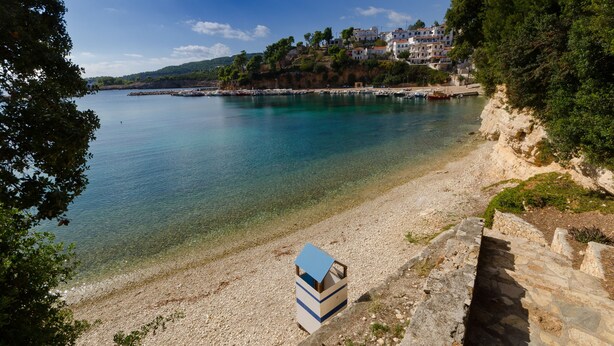A coastal area with a clear beach, a small building with a blue roof, and houses on a hillside.