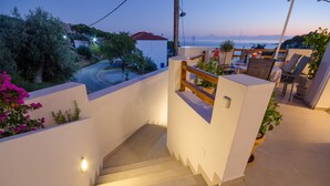 A staircase with a wooden handrail leading to a balcony with a view of the sea and a white building in the background.