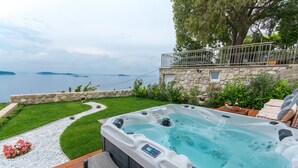A hot tub with a control panel, surrounded by a stone wall, a wooden deck, and a view of the sea.