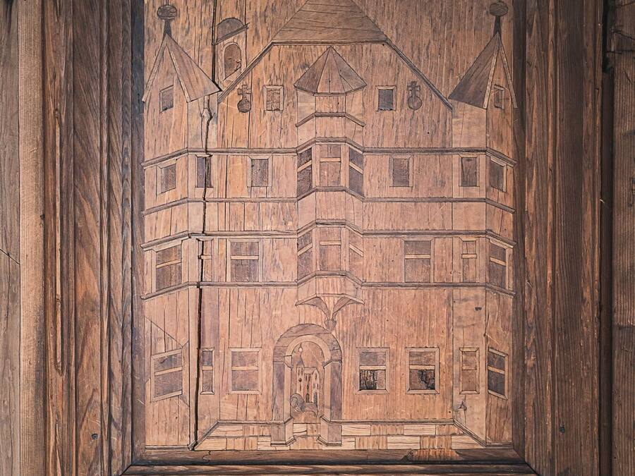 Un panel de madera con un diseño arquitectónico tallado, que incluye un edificio con varias ventanas y un arco central.