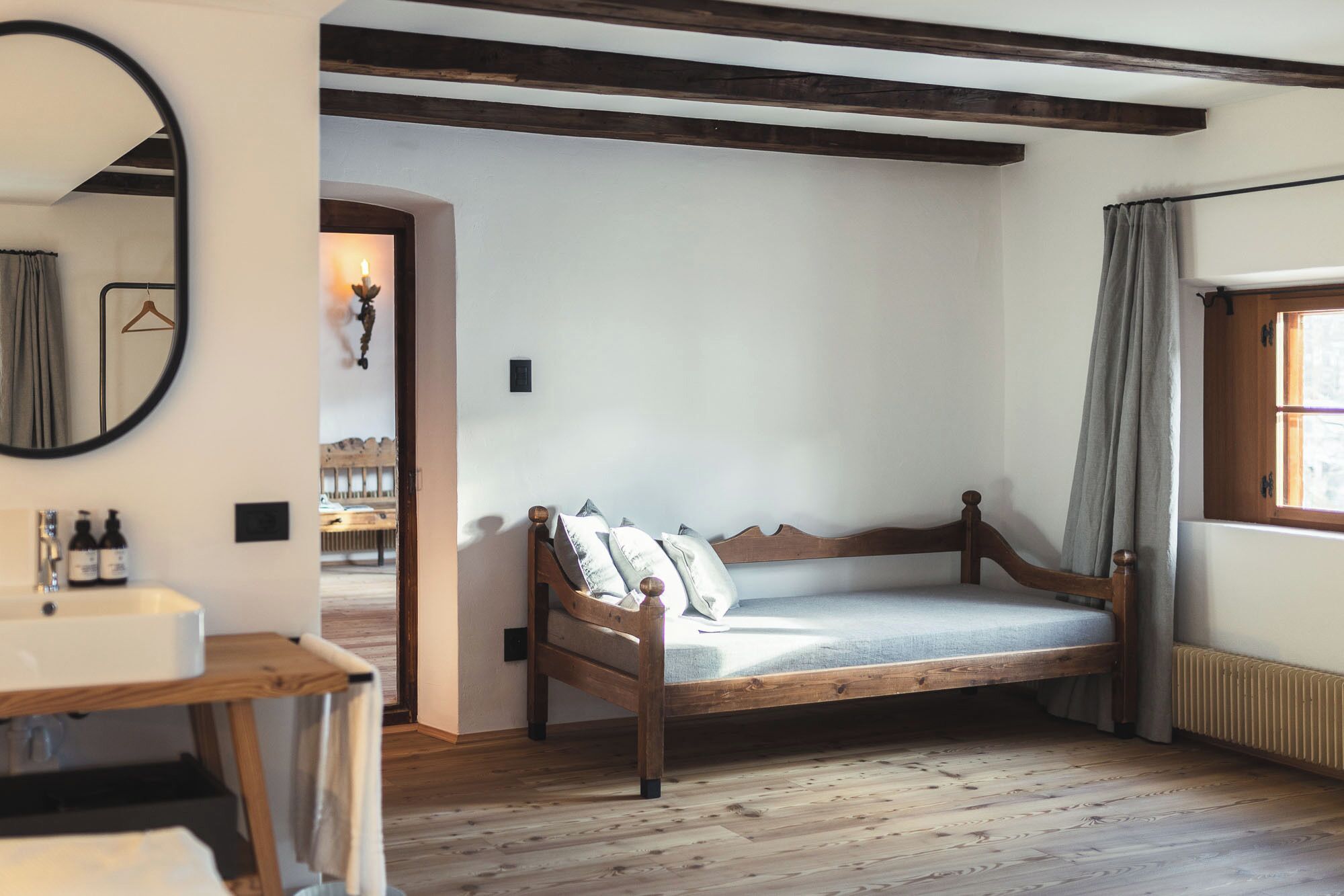 Quarto com cama de madeira, espelho redondo, mesa de cabeceira e janela com cortinas.