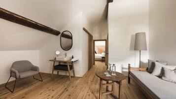 A room with a wooden floor, a sofa, a small table, a chair, a bedside table, a lamp, and a mirror.
