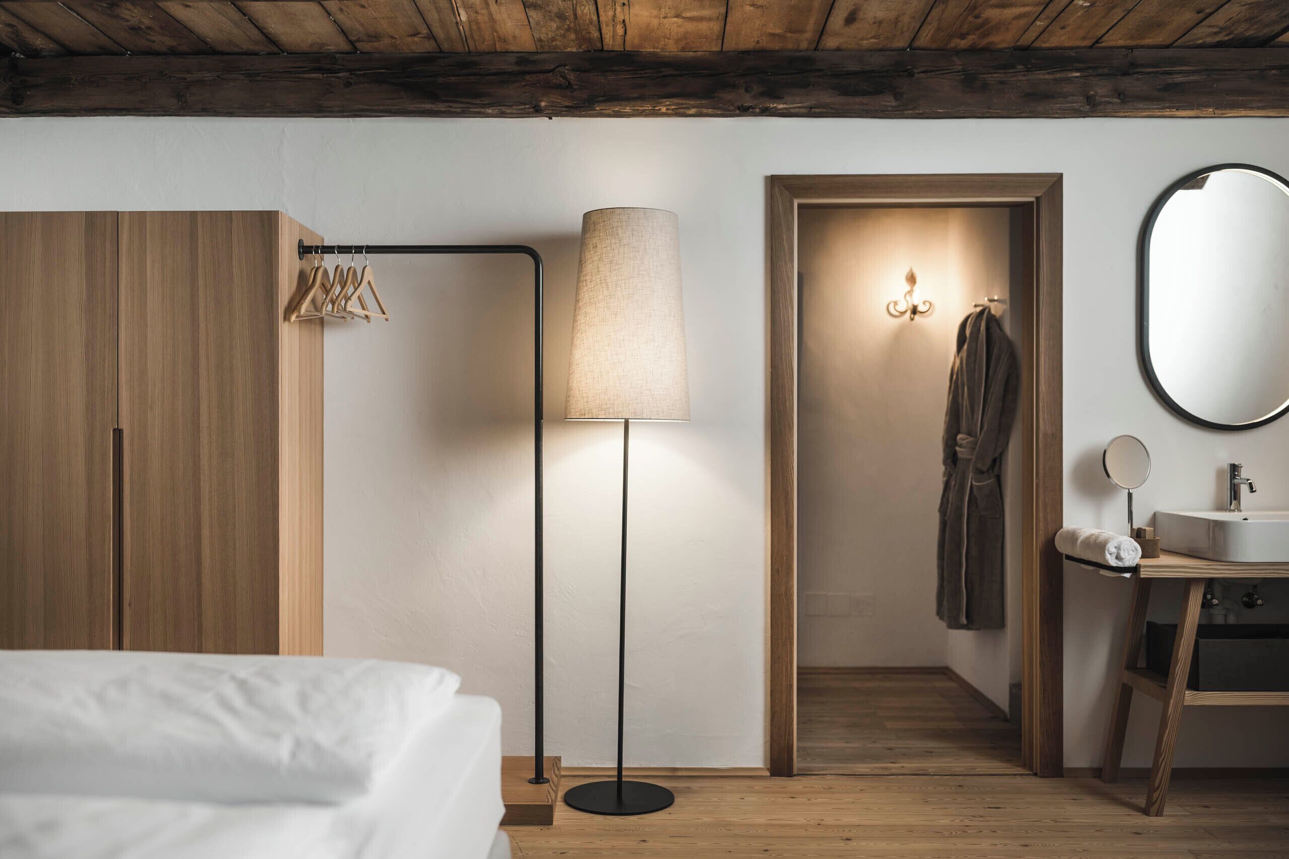 Quarto com cama, luminária de chão, guarda-roupa, espelho e banheiro com pia e porta-toalhas.