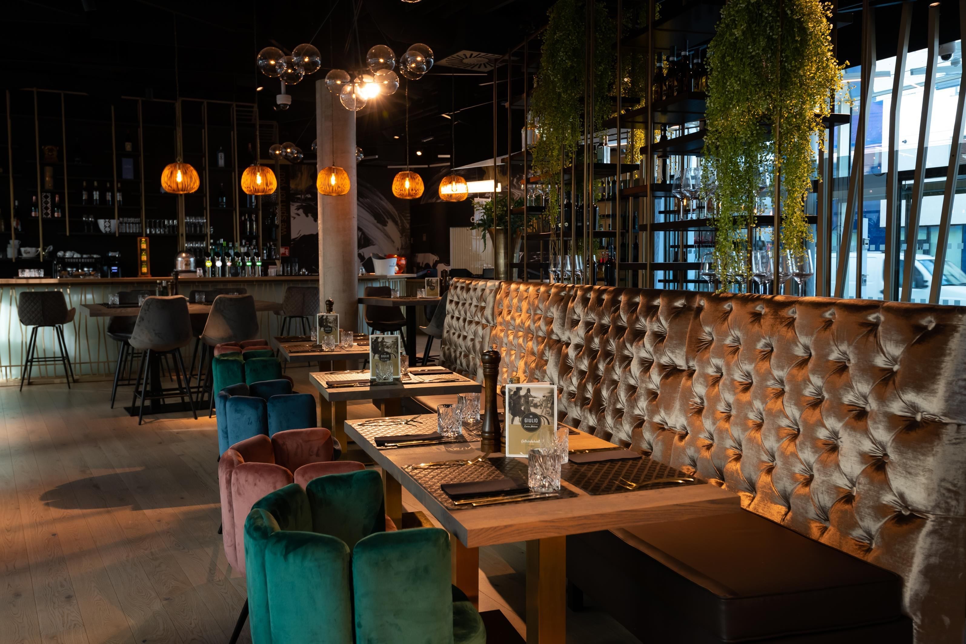 A restaurant interior with a bar area, seating with cushioned chairs, and pendant lighting.