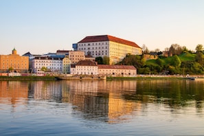 A riverside view of a historic building complex with multiple buildings, including a prominent white structure with a red roof. - Best Western Hotel Spinnerei Linz (Linz)
