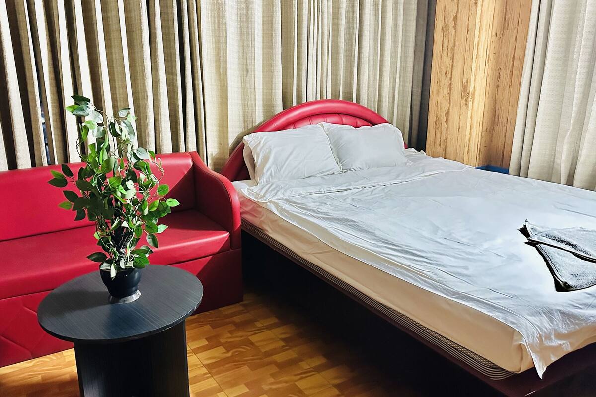 A hotel room with a red sofa, a small round table with a plant, a bed with white bedding, and wooden paneling on the walls.