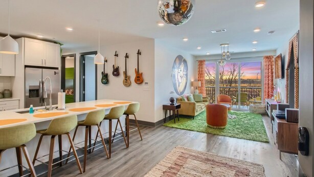 A modern kitchen with a bar area, a dining space with chairs, and a living area with a sofa and a view of the sunset.