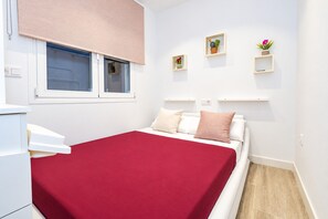 A bedroom with a bed, a window with blinds, a bedside table, and a wall-mounted shelf with decorative items.