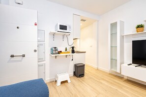 A modern, minimalistic room with a bed, a microwave, a small stool, a trash bin, and a TV.