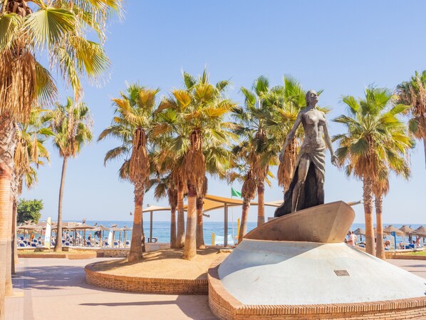 A beachside area with palm trees, a statue, and a sandy area with a circular brick structure. - Cubo's Apartment Front Line Beach Fuengirola / First line of beach / Air conditioning (Fuengirola)
