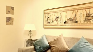 Un salon avec un canapé beige, deux coussins, un petit pouf rond bleu, une lampe fixée au mur et une œuvre d’art encadrée représentant une scène historique.