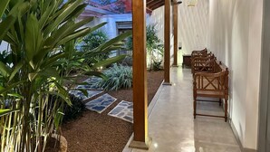 A covered outdoor area with wooden pillars, a row of rattan chairs, and a stone pathway leading to a garden with various plants.